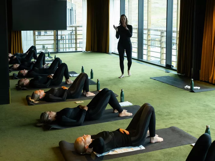 Damer som ligger på et grønt gulv, og gjør yoga, samt en instruktør som står å snakker.