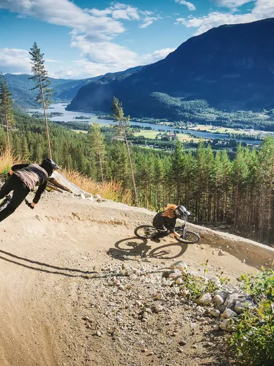 Den Ravinde i Nesbyen Foto Lars Storheim, to personer som sykler og i bakgrunnen er det fjell og skog.