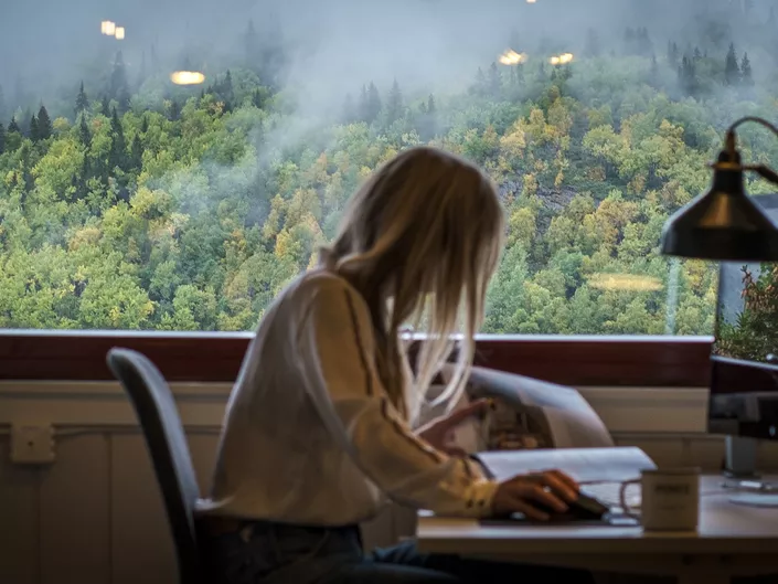 Dame som leser i et blad, foran en kontorpult. I bakgrunnen er et stort vindu.