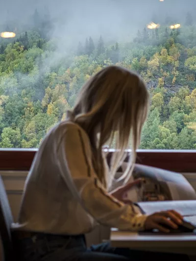 Dame som leser i et blad, foran en kontorpult. I bakgrunnen er et stort vindu.