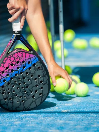 Bildet er av en hånd som plukker opp padel-baller med den ene håden, og holder rekkerten i den andre.