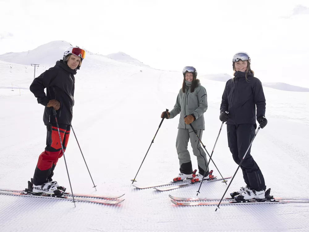 To damer og en mann som står i skibakken og ser inn i kameraet, ikledd skiklær.