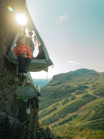 Mann som sitter med beina ut av skysleeping teltet, mens en dame ligger med hodet ut. I bakgrunnen av teltet er det flere andre telt og fjell.