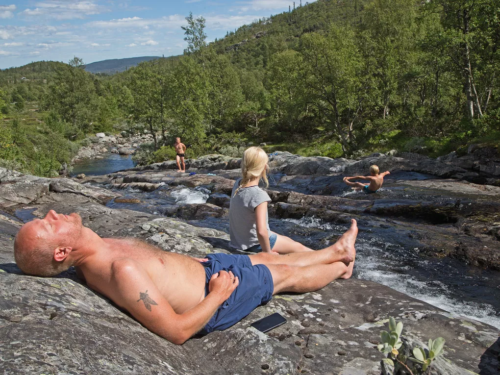 Mann soler seg, imens barna leker i elva.