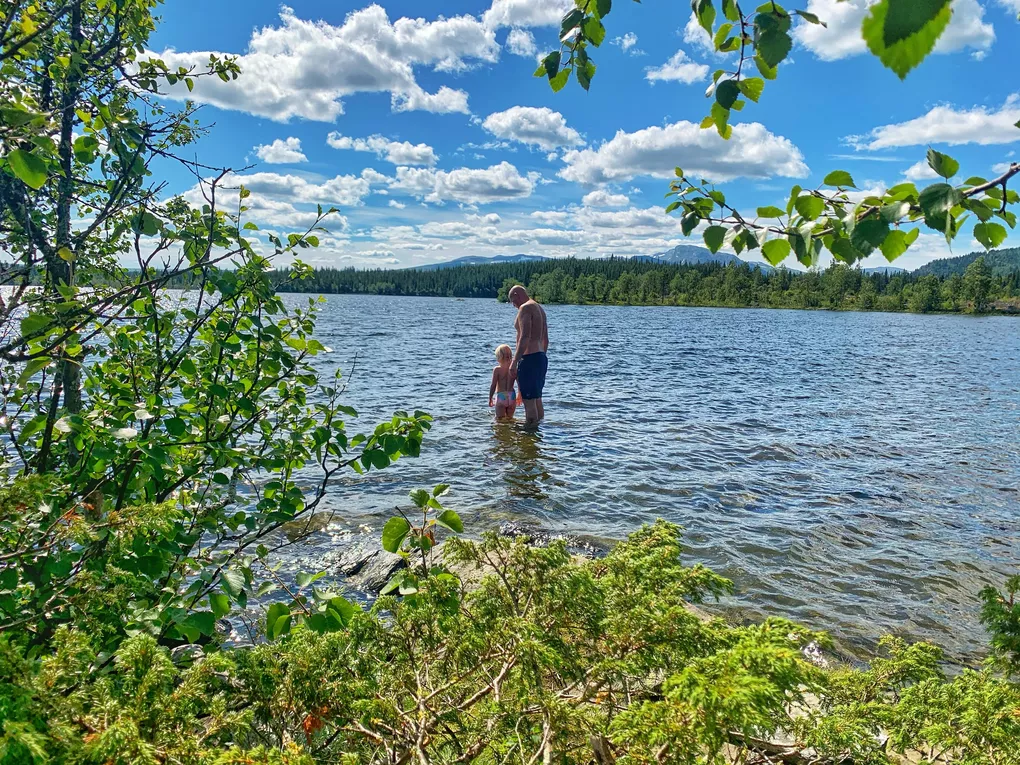 Far og datter på vei ut for å bade, det er sol og fint vær. I bakgrunnen er det grønne blader og trær.