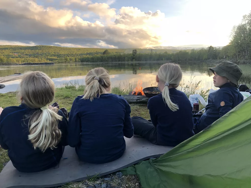 Barn som telter og sitter ved et bål. I bakgrunnen er det et vann.