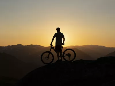 Mann og sykkel som står på toppen av et fjell i solnedgangen