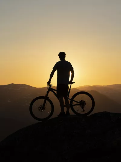Mann og sykkel som står på toppen av et fjell i solnedgangen